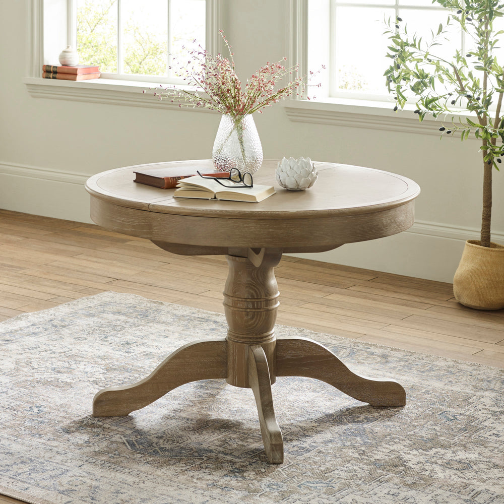 Round wooden table with light brown finish, pedestal base with four feet, vase and books on top, by window