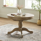 Round wooden table with light brown finish, pedestal base with four feet, vase and books on top, by window