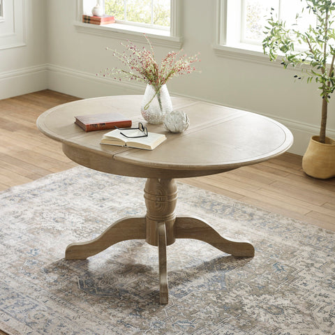 Round wooden table with light brown finish near windows, decorative items on top, floral rug underneath