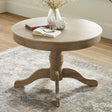 Round wooden table with light brown finish, pedestal base, books, glasses, and vase on top