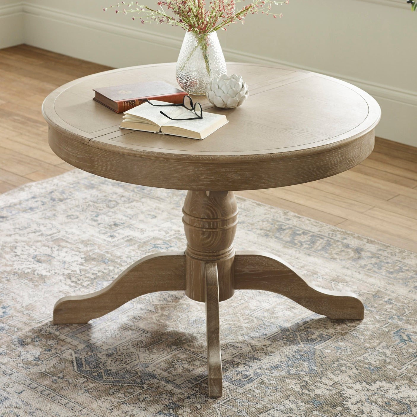 Round wooden table with light brown finish, pedestal base, books, glasses, and vase on top