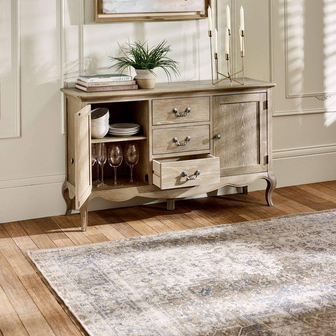 The Camille French Style 2 Door 2 Drawer Sideboard in limed oak features a light, weathered finish with elegant curved legs, showcasing two drawers and two doors that reveal ample storage space, set against a warm wooden floor and complemented by decorative items and a soft area rug