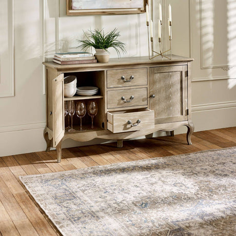 The Camille French Style 2 Door 2 Drawer Sideboard in limed oak features a light, weathered finish with elegant curved legs, showcasing two drawers and two doors that reveal ample storage space, set against a warm wooden floor and complemented by decorative items and a soft area rug