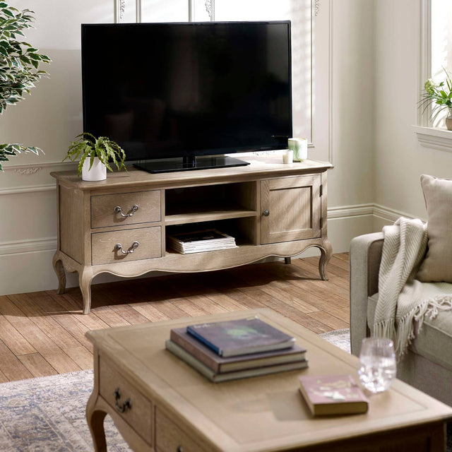 The Camille French Style TV Unit in limed oak features elegant curves, two drawers, and an open shelf, beautifully complementing a light-filled living room setting with a cozy sofa and decorative plants
