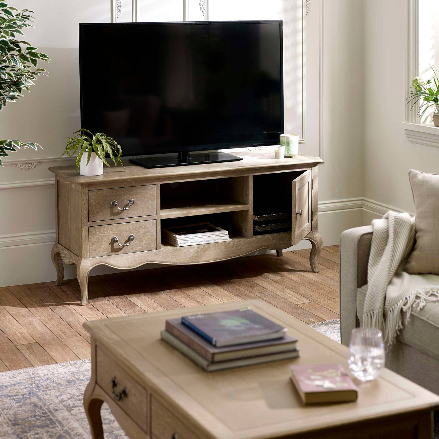 The Camille French Style TV Unit in limed oak features elegant curved legs, two drawers, and open shelving, beautifully complementing a light-filled living room setting with a cozy sofa and decorative plants