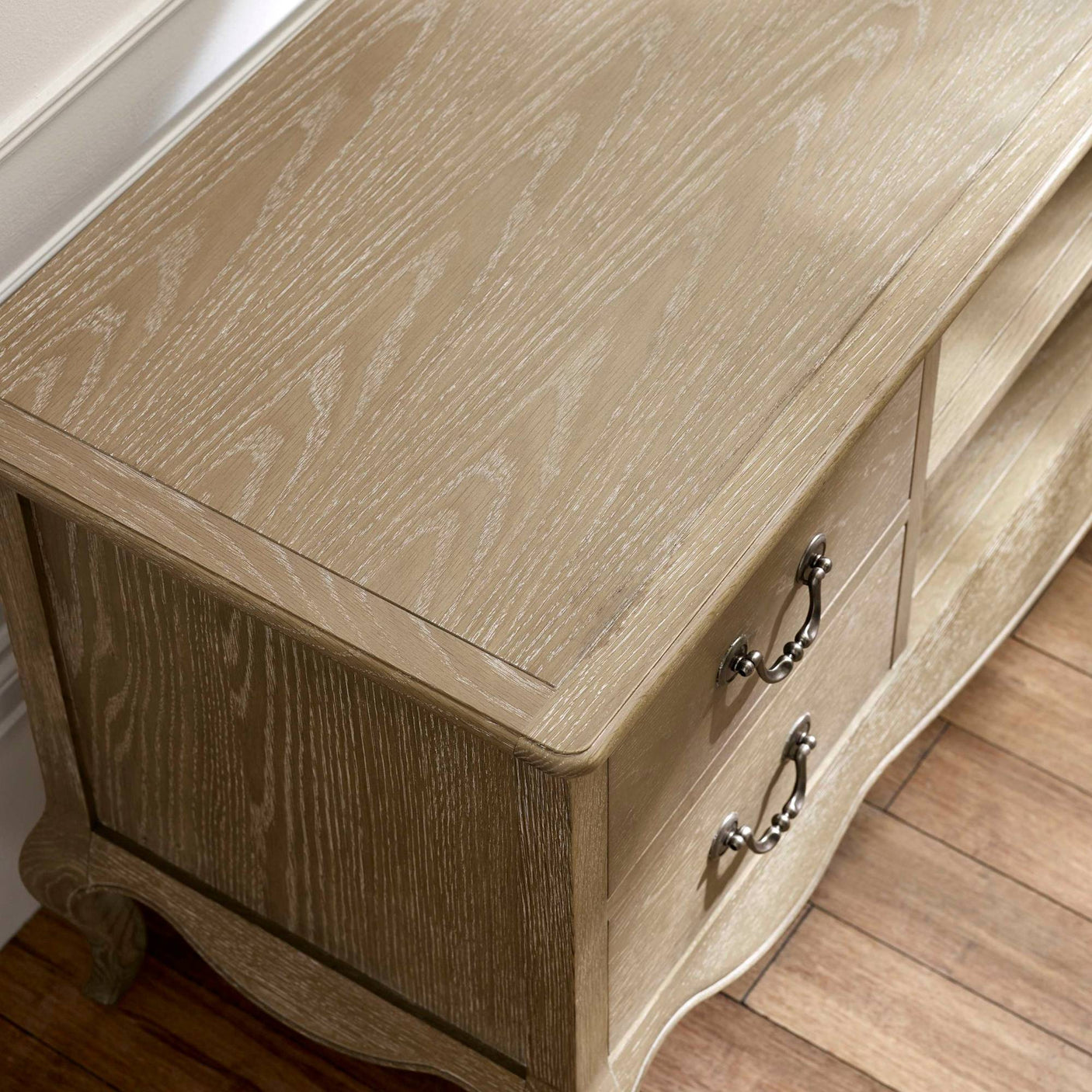 The Camille French Style TV Unit in limed oak features a smooth, textured surface with elegant detailing and two drawers, set against a warm wooden floor