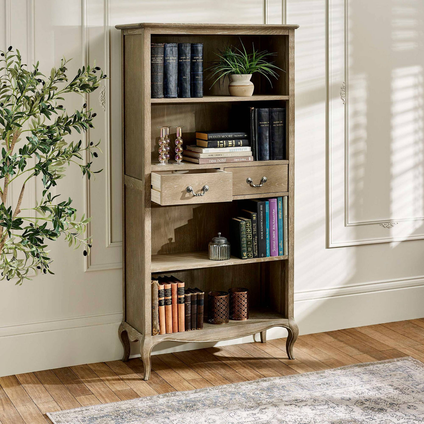 The Camille French Style Tall Bookcase with Drawers in limed oak features elegantly curved legs, two spacious drawers, and multiple shelves adorned with books and decorative items, set against a softly lit room with a patterned rug and a decorative plant