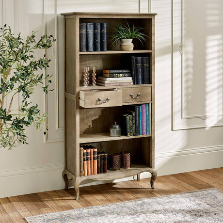 The Camille French Style Tall Bookcase with Drawers in limed oak features elegantly curved legs, two spacious drawers, and multiple shelves adorned with books and decorative items, set against a softly lit room with a patterned rug and a decorative plant