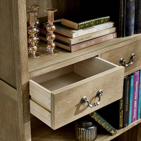 The Camille French Style Tall Bookcase with Drawers features a limed oak finish, showcasing a spacious design with open shelves filled with books and decorative candlesticks, complemented by elegantly styled drawers with metal handles