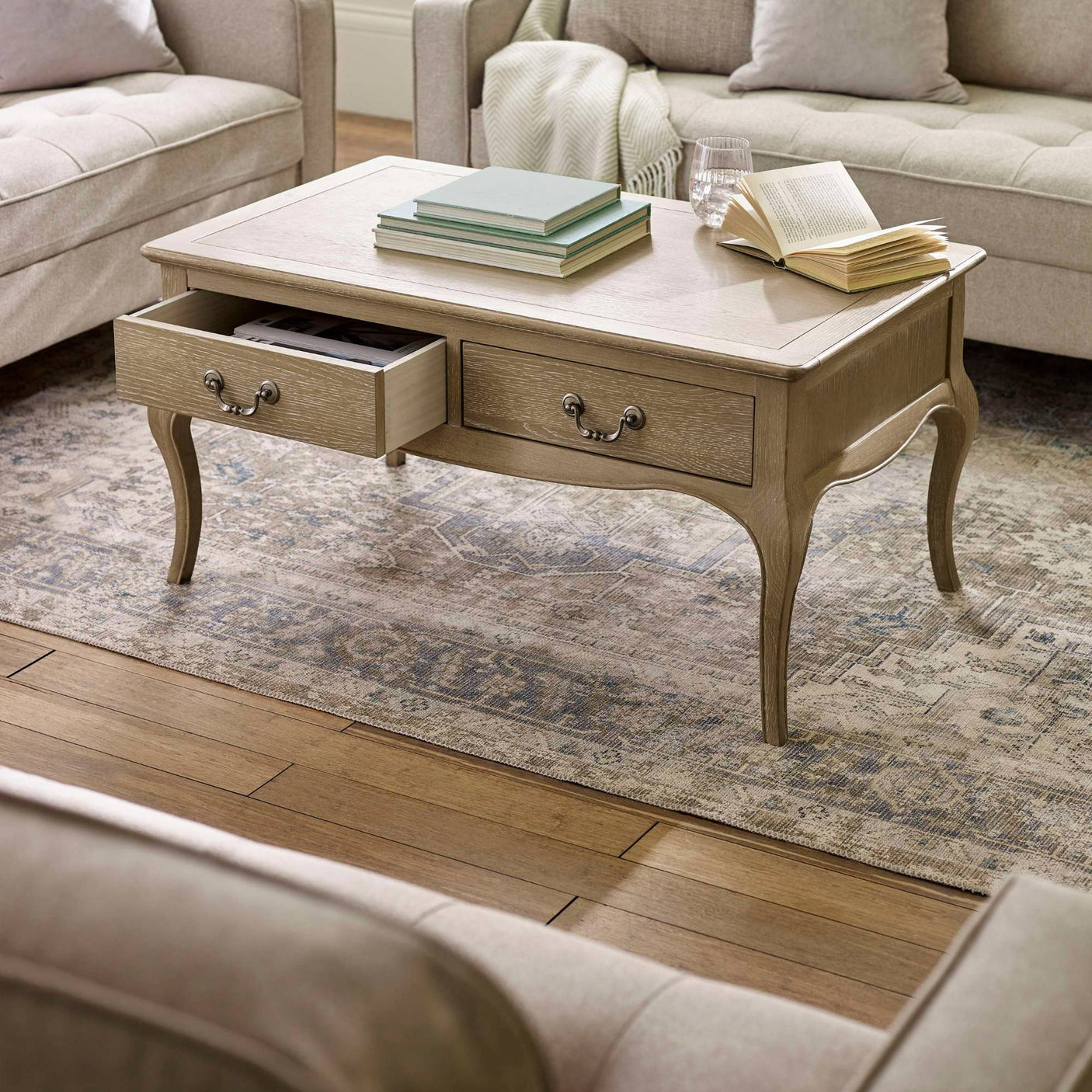 The Camille French Style Rectangular Coffee Table in limed oak features a smooth top, two spacious drawers with decorative handles, and elegantly curved legs, set against a cozy living room backdrop with neutral-toned sofas and a patterned area rug