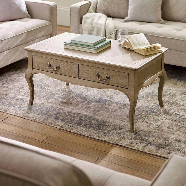 The Camille French Style Rectangular Coffee Table in limed oak features elegant curved legs and two drawers, beautifully set on a patterned area rug in a cozy living room