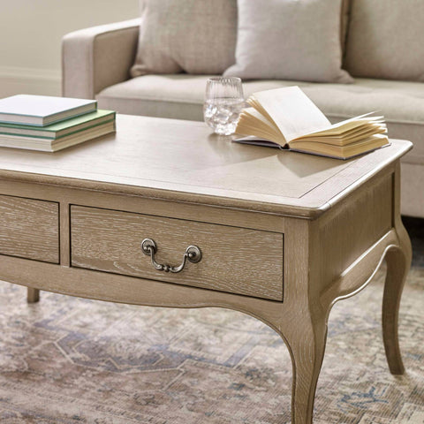 The Camille French Style Rectangular Coffee Table in limed oak features a smooth surface with two drawers and elegant curved legs, set in a cozy living room beside a light-colored sofa and decorative books