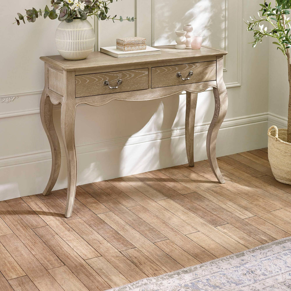 The Camille Console Table in limed oak features a classic design with elegantly curved legs, two drawers with decorative metal handles, and is set against a softly lit room with a neutral color palette and decorative accents