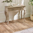 The Camille Console Table in limed oak features a classic design with elegantly curved legs, two drawers with decorative metal handles, and is set against a softly lit room with a neutral color palette and decorative accents