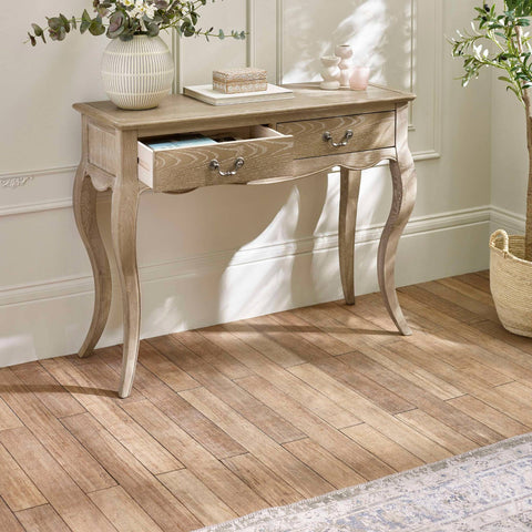 The Camille Console Table in limed oak features elegant curved legs and two drawers, set against a softly lit room with a neutral color palette and decorative accents