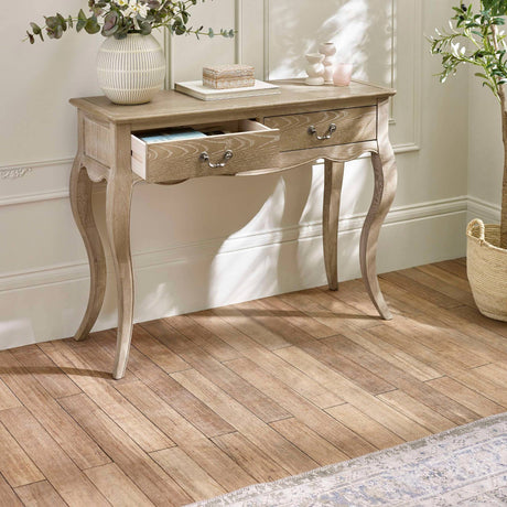 The Camille Console Table in limed oak features elegant curved legs and two drawers, set against a softly lit room with a neutral color palette and decorative accents