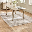 The Camille French Style Rectangular 4-6 Seater Extending Dining Table in limed oak features elegantly curved legs and a smooth surface, set on a light wooden floor with a decorative area rug and a vase of flowers, creating a warm and inviting dining space