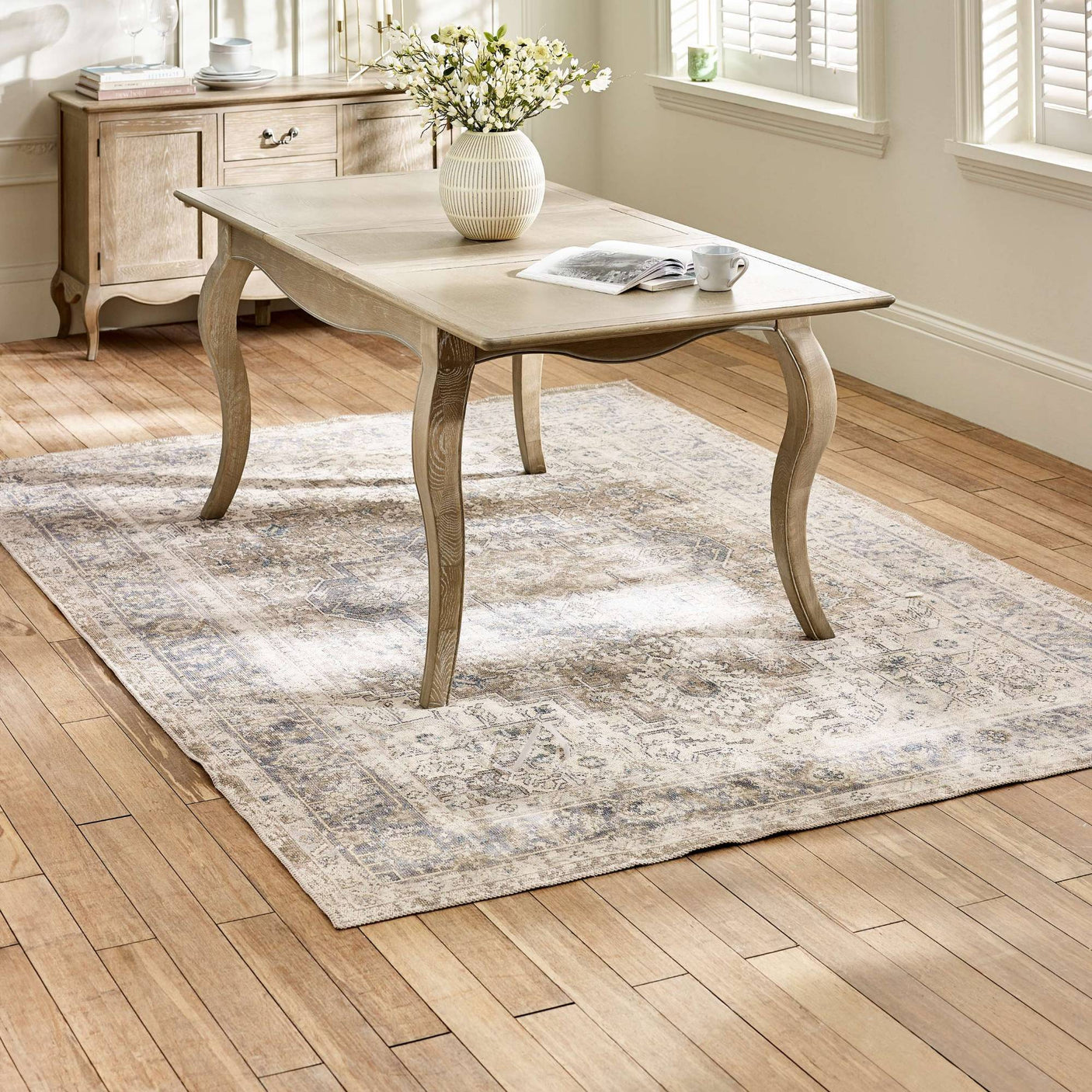 The Camille French Style Rectangular 4-6 Seater Extending Dining Table in limed oak features elegantly curved legs and a smooth surface, set on a light wooden floor with a decorative area rug and a vase of flowers, creating a warm and inviting dining space
