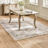 The Camille French Style Rectangular 4-6 Seater Extending Dining Table in limed oak features elegantly curved legs and a smooth surface, set on a light wooden floor with a decorative area rug and a vase of flowers, creating a warm and inviting dining space