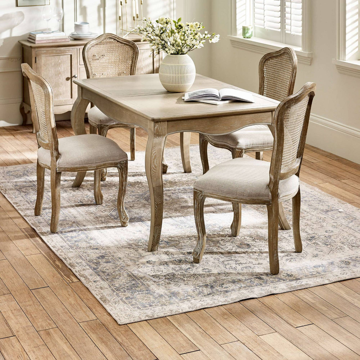The Camille French Style Rectangular 4-6 Seater Extending Dining Table in limed oak features elegantly curved legs and a smooth surface, surrounded by upholstered chairs with caned backs, all set on a patterned rug in a bright, airy dining room