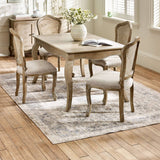 The Camille French Style Rectangular 4-6 Seater Extending Dining Table in limed oak features elegantly curved legs and a smooth surface, surrounded by upholstered chairs with caned backs, all set on a patterned rug in a bright, airy dining room