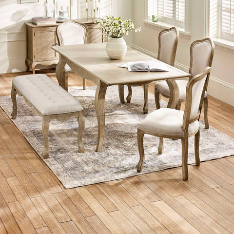 The Camille French Style Rectangular 4-6 Seater Extending Dining Table in limed oak features elegantly curved legs and is paired with upholstered chairs and a tufted bench, all set on a patterned rug in a bright, airy dining room