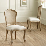 Two beige rattan dining chairs with carved light wood frames on light wood floor near white wall and window