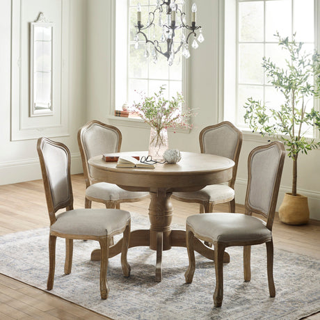 Round wooden dining table with four beige upholstered dining chairs in a bright, classic room setting.