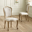 Two beige upholstered wooden dining chairs with carved legs on hardwood floor near paneled wall.