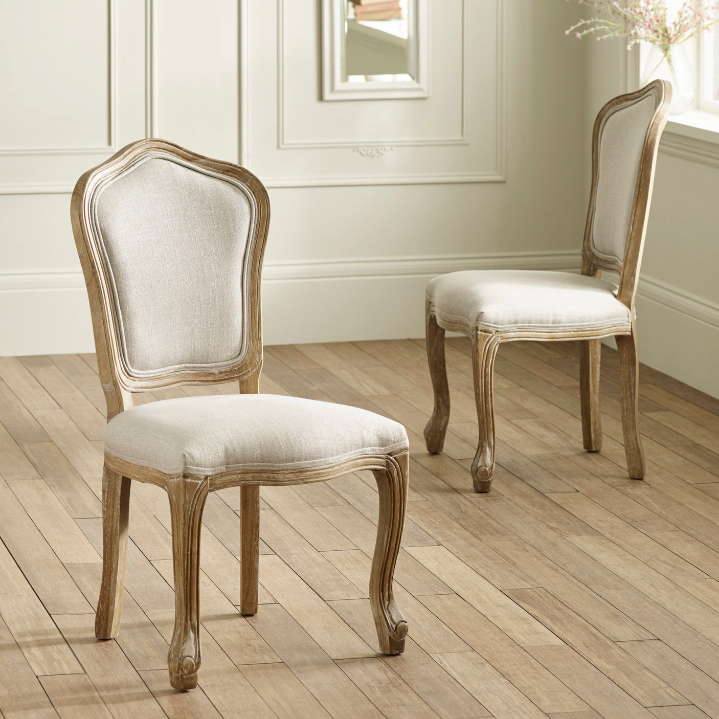 Two beige upholstered wooden dining chairs with carved legs on hardwood floor near paneled wall.