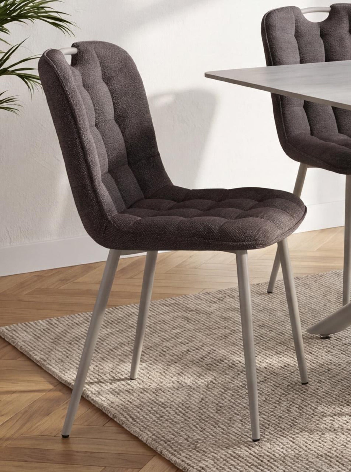 A dark brown tufted upholstered dining chair with slim silver metal legs, shown in a modern dining room setting next to a table on a textured rug.