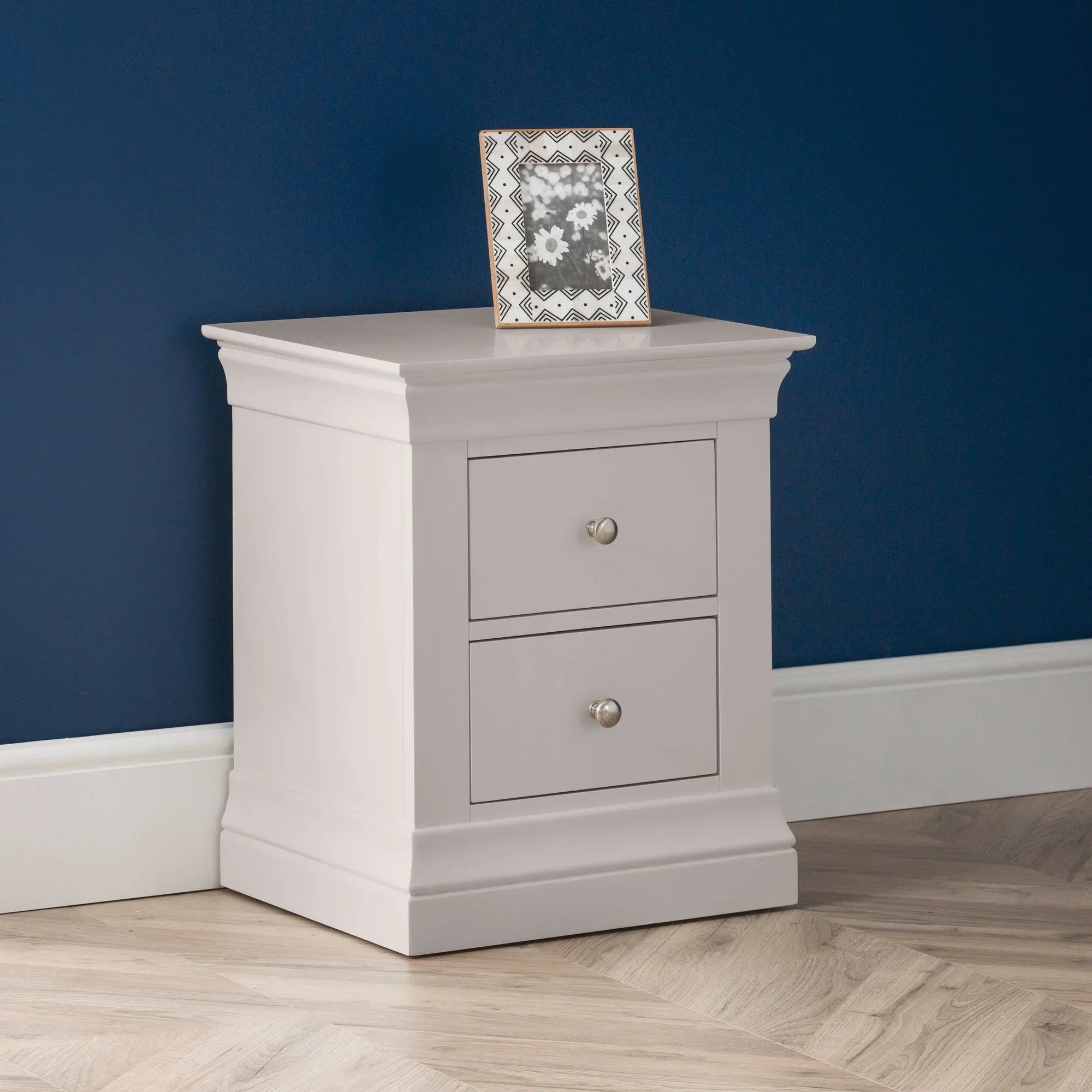Light grey two drawer bedside table with silver knobs on herringbone floor