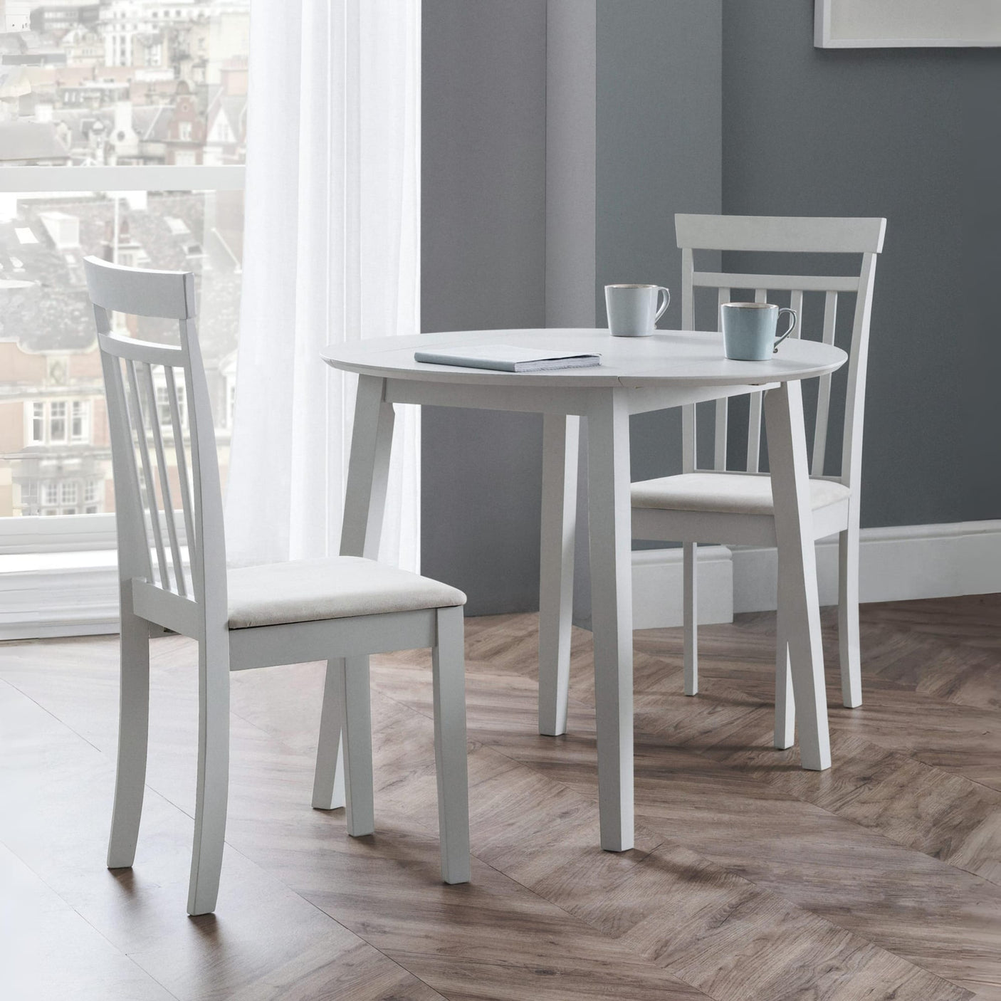 Round white dining table with two white chairs on wood floor by large window with city view and grey walls.