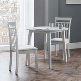 Round white dining table with two white chairs on wood floor by large window with city view and grey walls.