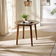 Round brown wooden table with angled legs on beige rug by window with potted plant and reading material