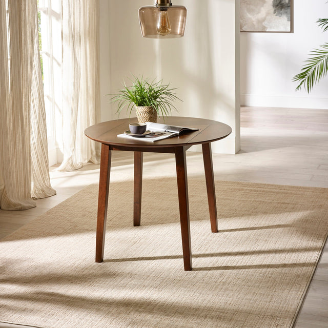 Round brown wooden table with angled legs on beige rug by window with potted plant and reading material