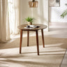 Round brown wooden table with angled legs on beige rug by window with potted plant and reading material