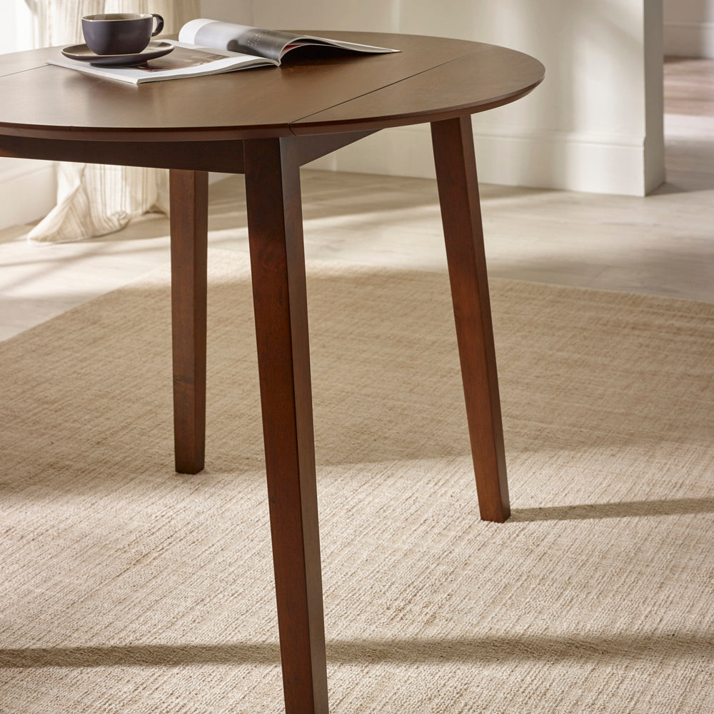 Side view of round brown wooden table with drop leaf folded down on beige rug with angled legs visible