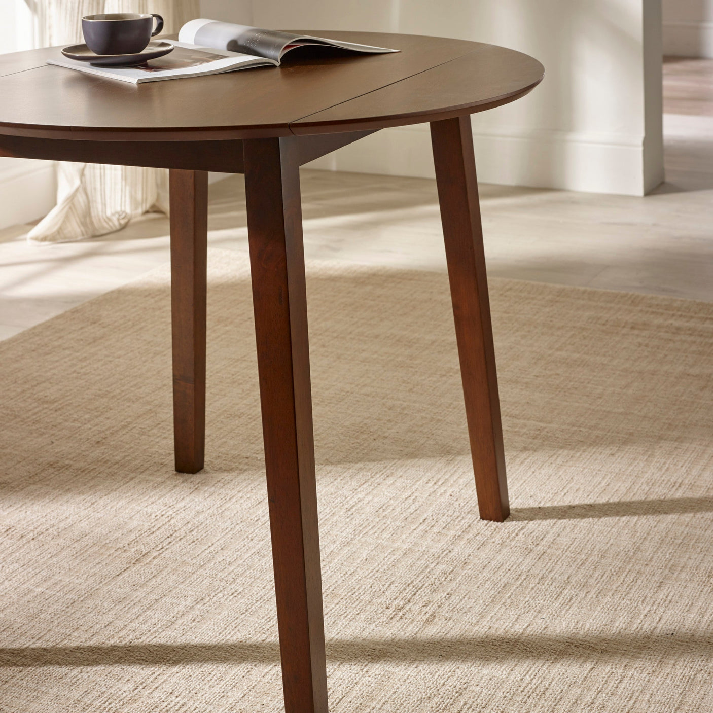 Side view of round brown wooden table with drop leaf folded down on beige rug with angled legs visible