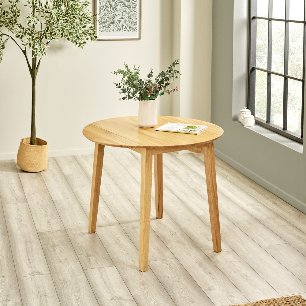Natural wood round dropleaf table with vase and magazine on top, sitting in bright airy room with plant
