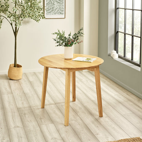 Natural wood round dropleaf table with vase and magazine on top, sitting in bright airy room with plant