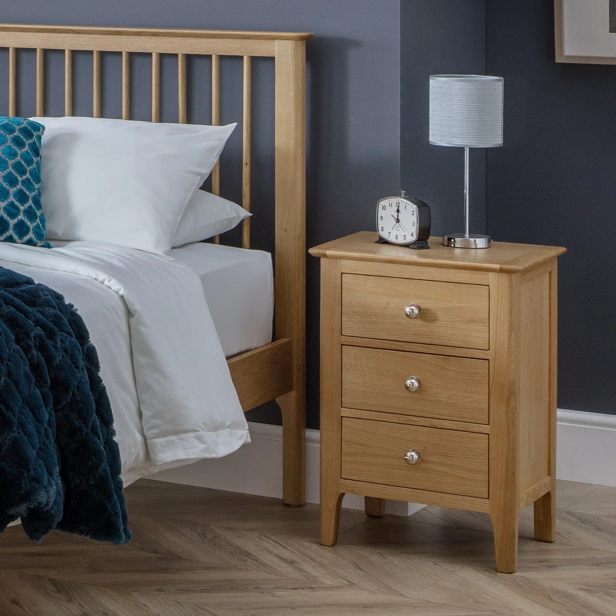 Oak three drawer bedside table beside bed with lamp and clock on top