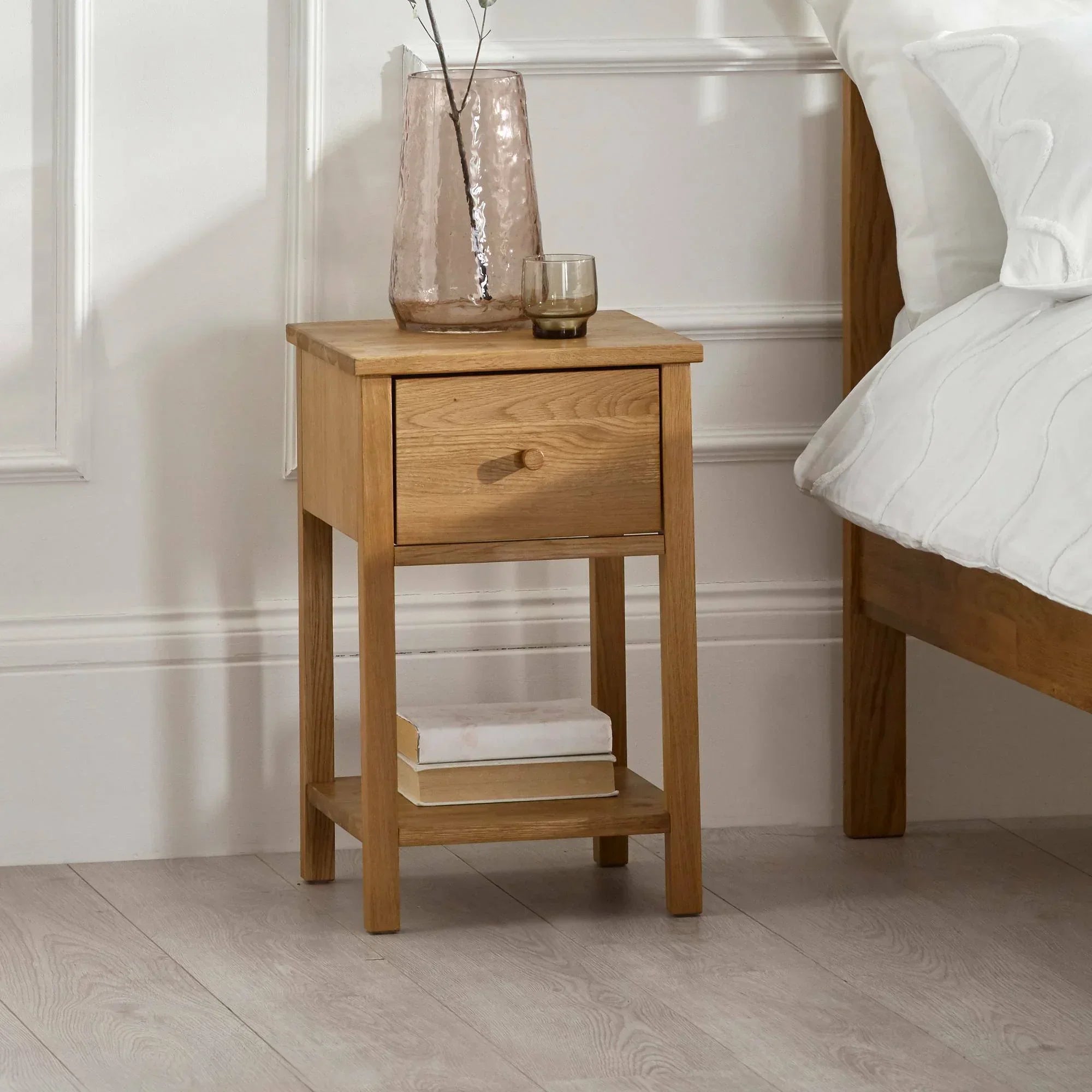 Oak bedside table with single drawer and shelf beside bed, vase and glass on top