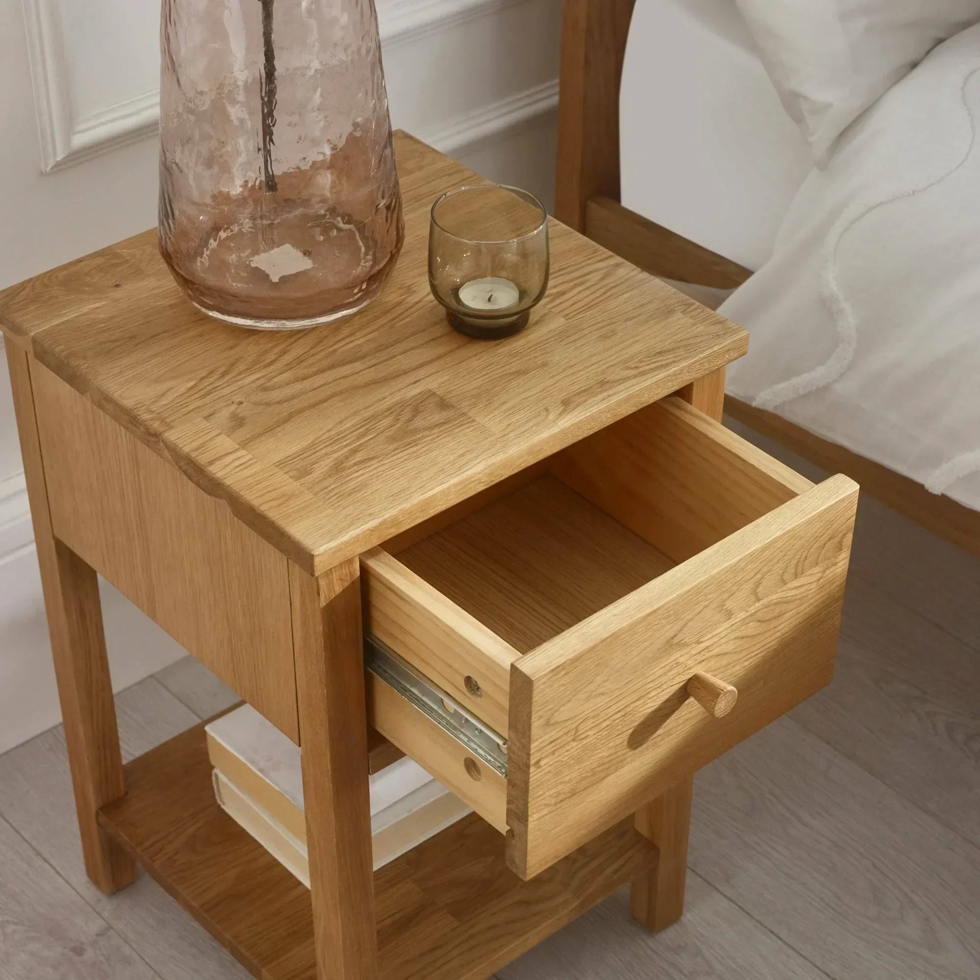 Oak bedside table with drawer open, glass vase and candle on top, books on lower shelf