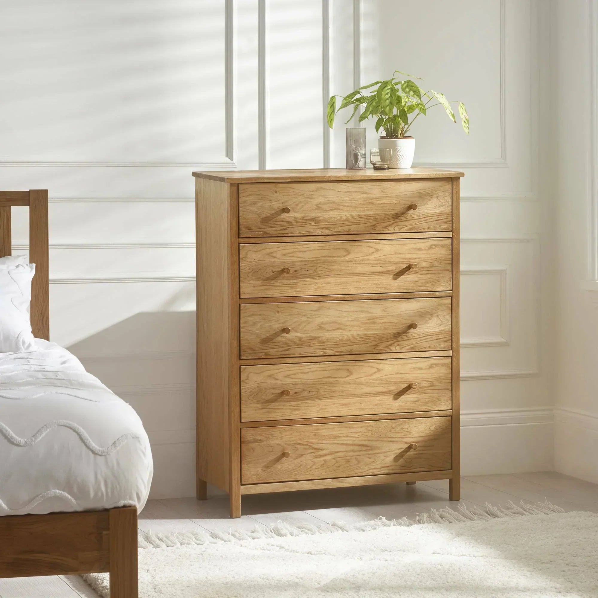 Oak chest of drawers with five wide drawers in bright bedroom, plant on top