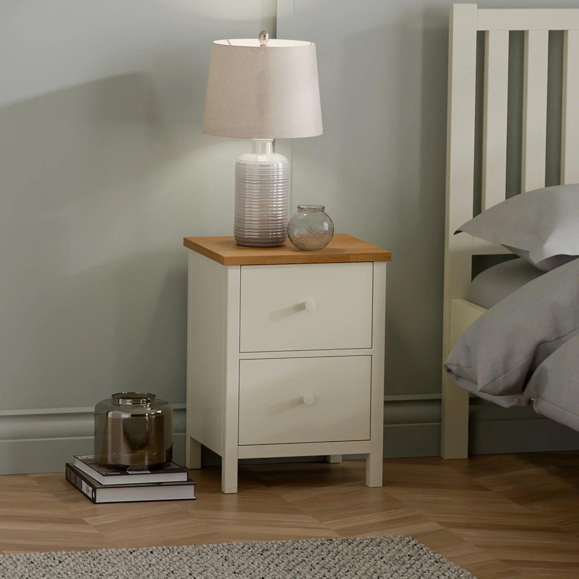 Ivory bedside table with two drawers and oak top beside bed, lamp and decor on surface