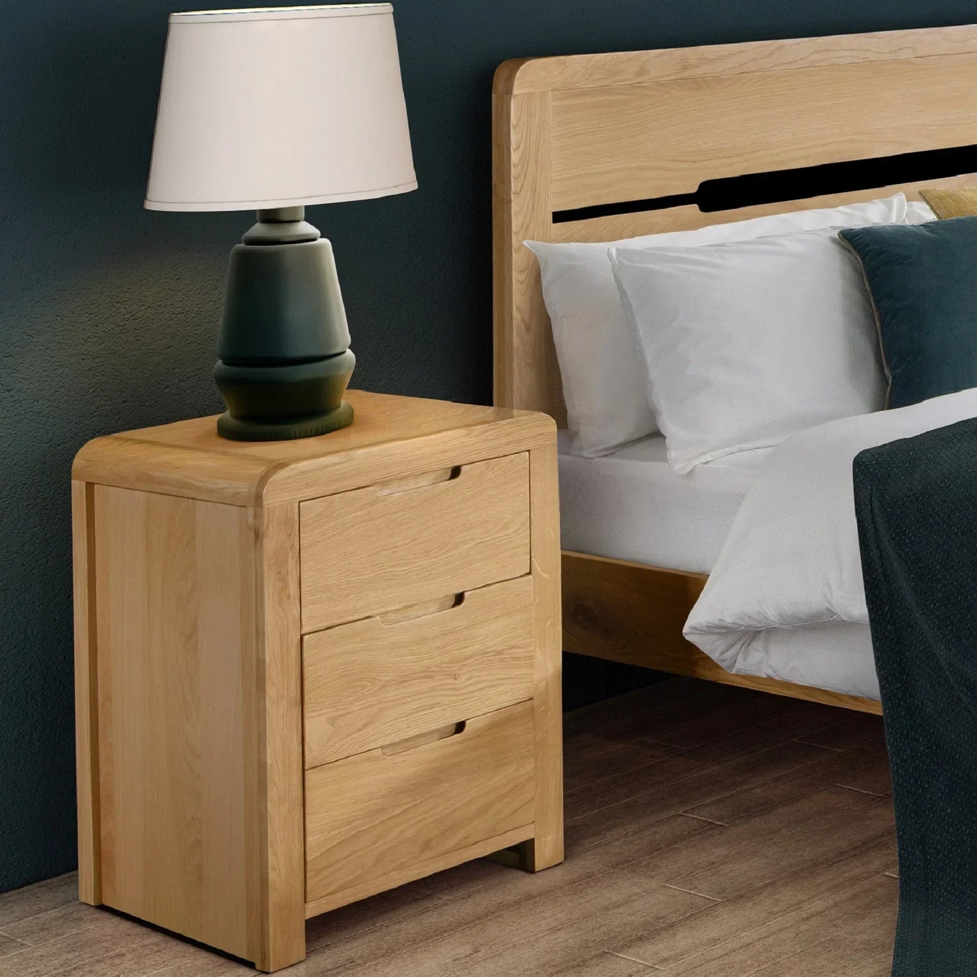 Oak bedside table with three drawers beside bed, lamp on top, wooden flooring visible
