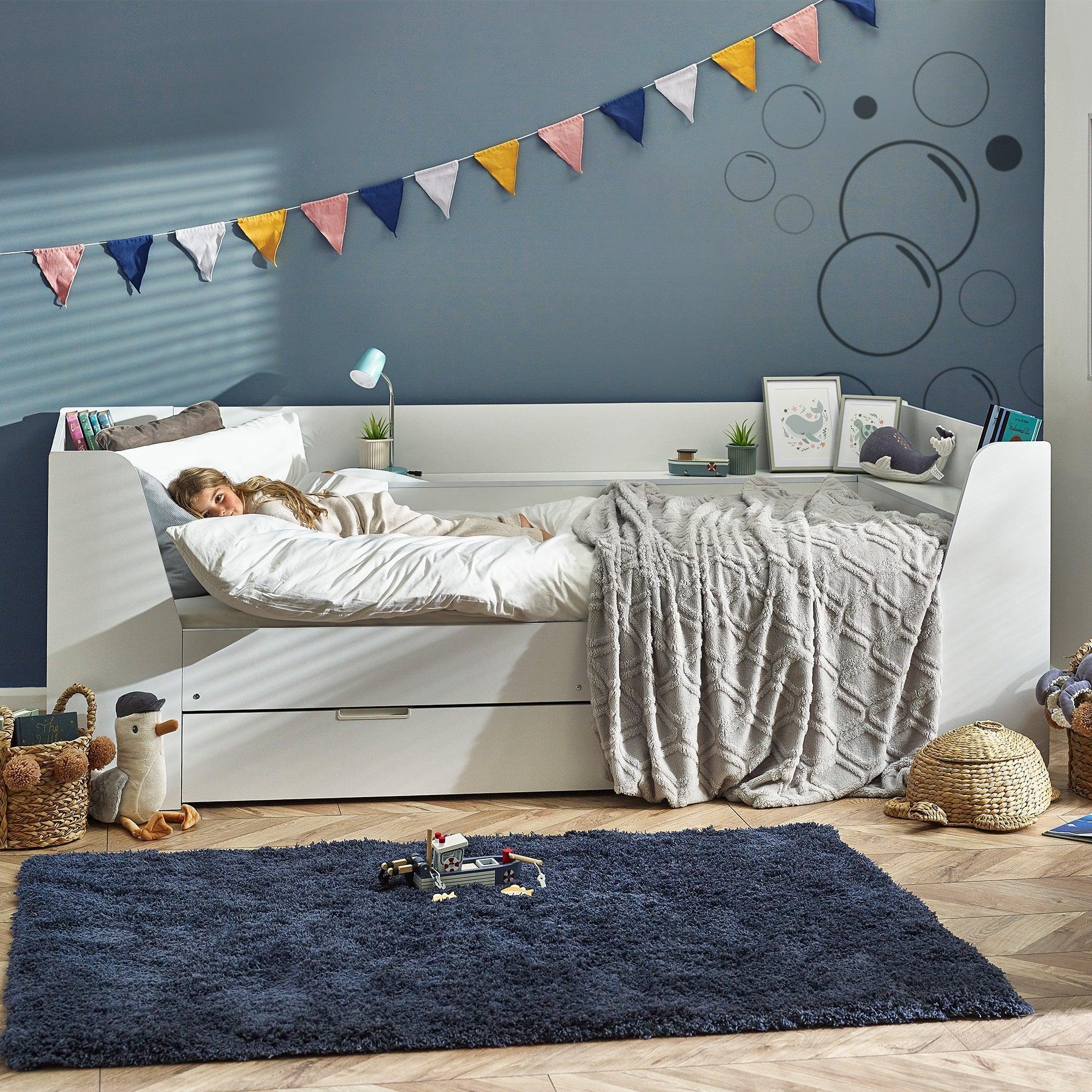 White daybed with storage drawer in modern kids room, child resting under duvet, decorative flags