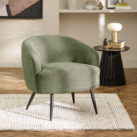 Green fabric barrel chair with black tapered legs on patterned rug next to black side table with lamp.