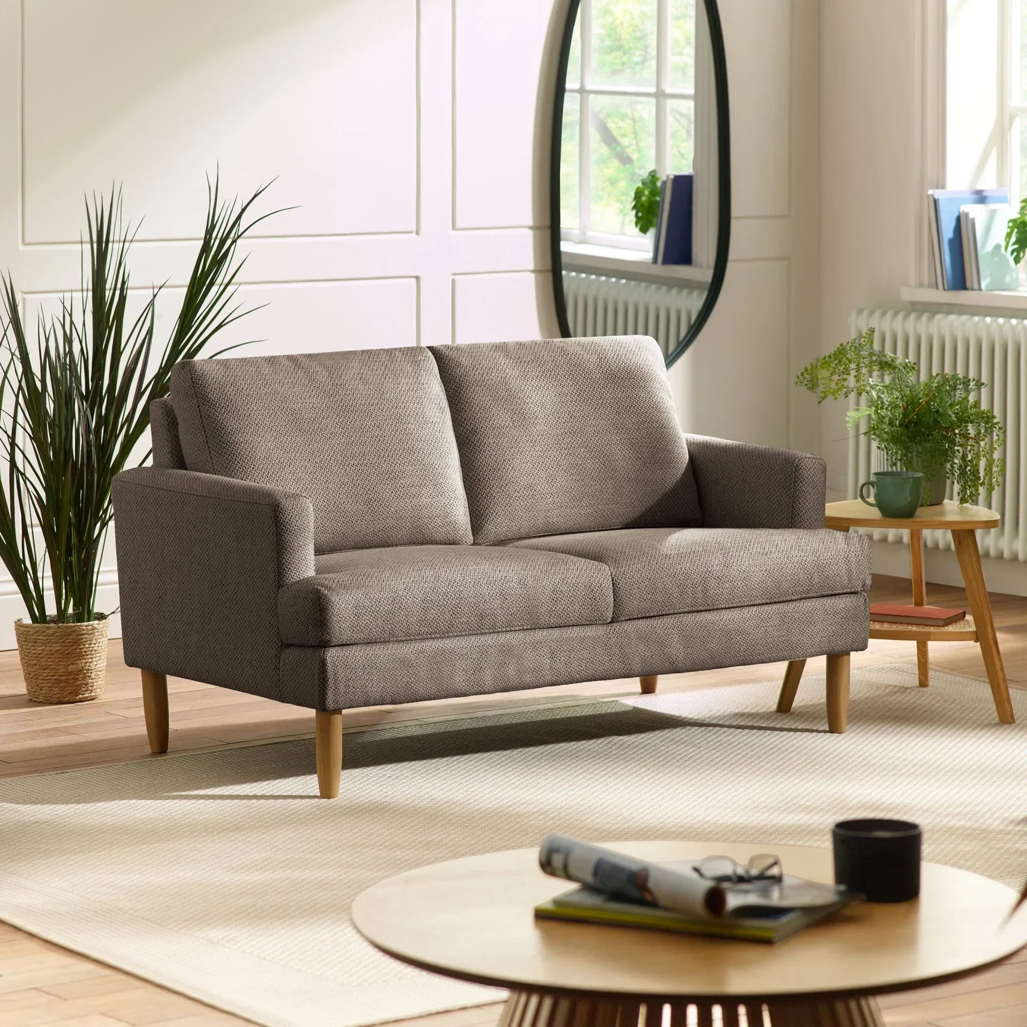 Grey fabric sofa with wooden legs in bright living room, plants and round table nearby